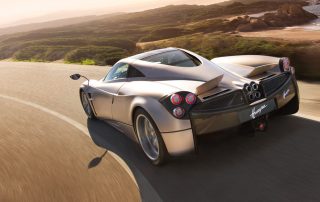The Pagani Huayra, silver, drives along a windy country road with the sun bearing down overhead.