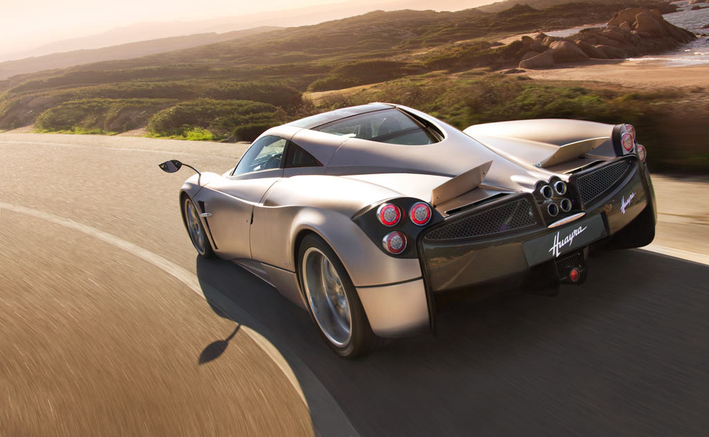 The Pagani Huayra, silver, drives along a windy country road with the sun bearing down overhead.