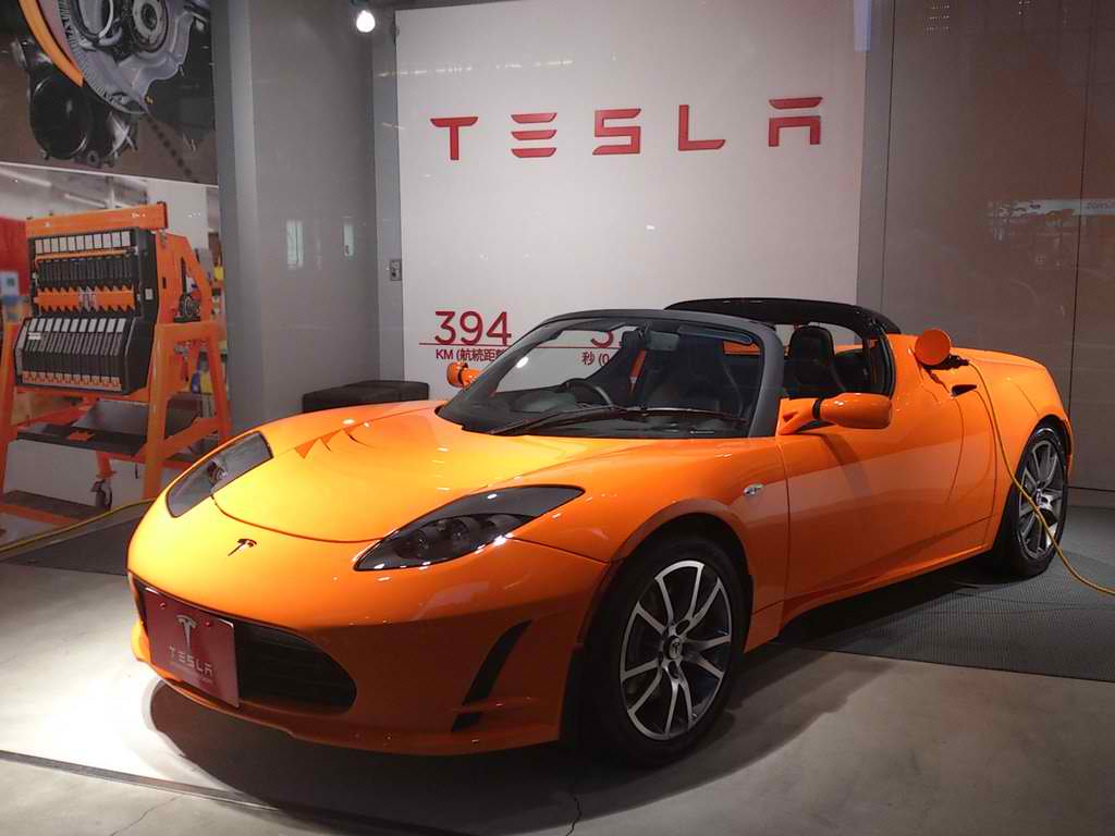 An orange Tesla Roadster is on display in front of a Tesla sign.