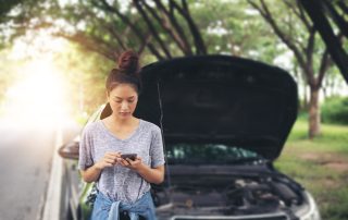 Girl standing in front of broken-down car texting for help.