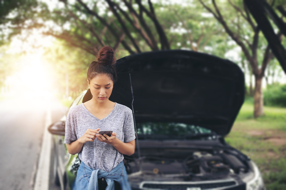 Girl standing in front of broken-down car texting for help.